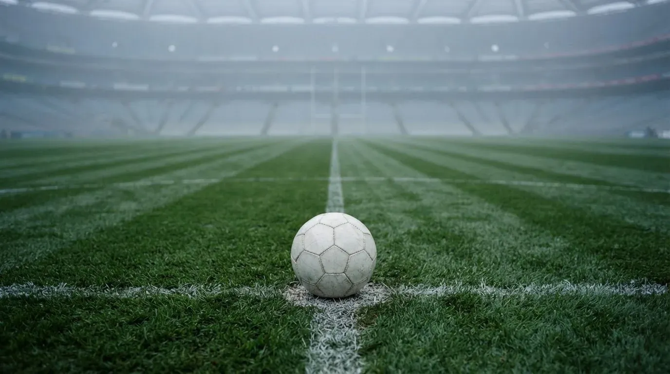 Ein Fußball liegt auf dem Mittelkreis eines leeren Rasenplatzes am frühen Morgen, im Hintergrund eine ferne Tribüne im Dunst.