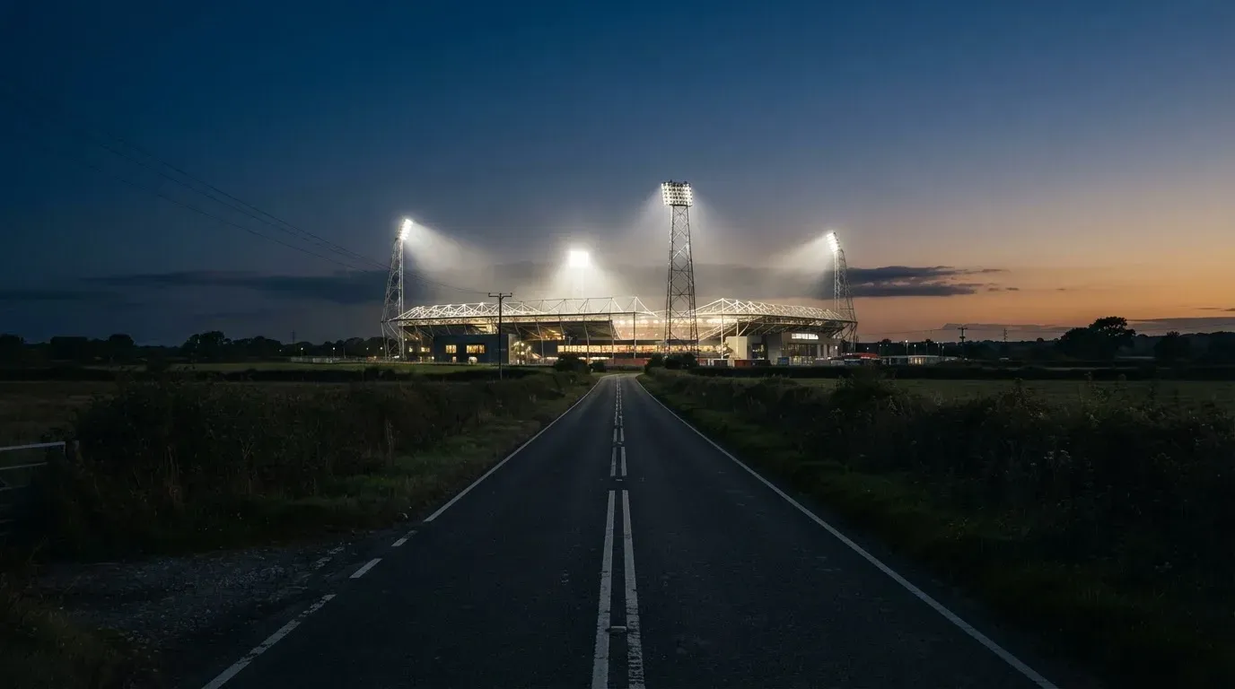 Ein Flutlichtstadion erhebt sich bei Dämmerung in der Ferne hinter einer leeren Landstraße, die Lichter sind eingeschaltet.