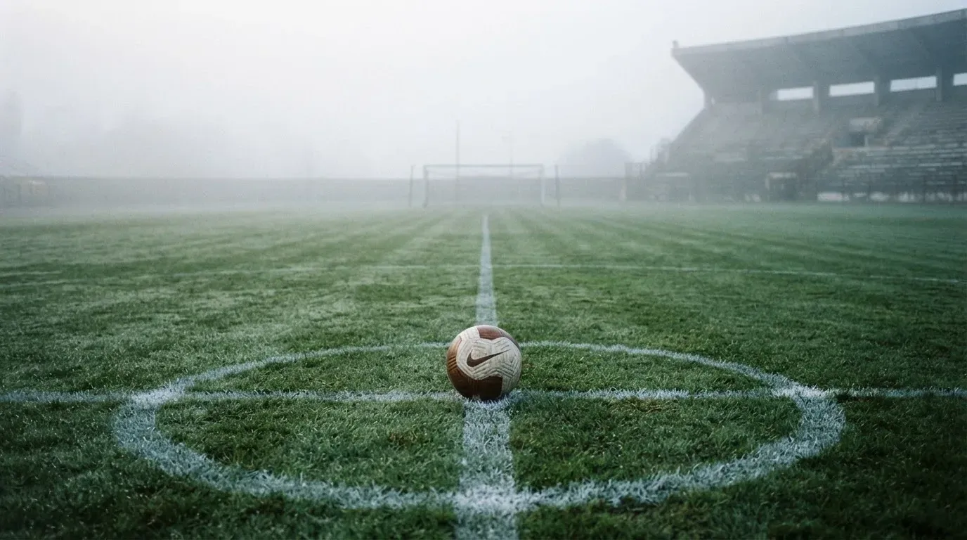 Ein Fußball liegt auf dem Anstoßpunkt eines leeren Rasenplatzes im Morgendunst, im Hintergrund eine ferne Tribüne.