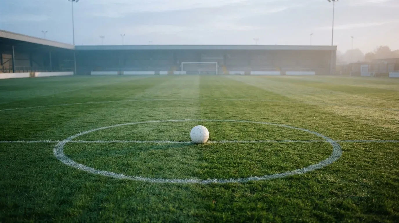 Ein Fußball liegt auf dem Mittelkreis eines leeren Rasenplatzes am frühen Morgen, im Hintergrund eine ferne Tribüne im Dunst.