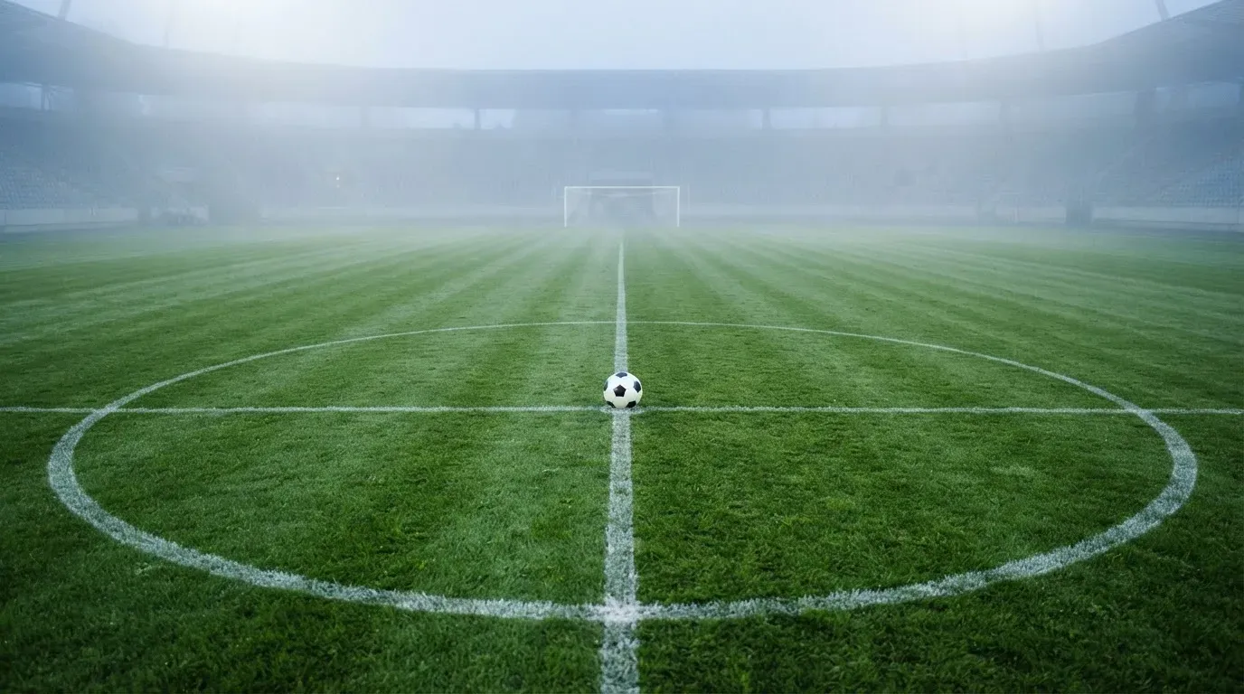 Ein Fußball liegt auf dem Anstoßpunkt eines leeren Rasenplatzes im Morgennebel.