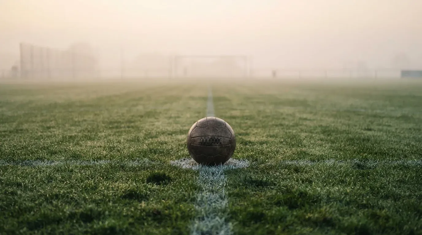 Ein Fußball liegt auf dem Anstoßpunkt eines leeren Rasenplatzes im Morgennebel.