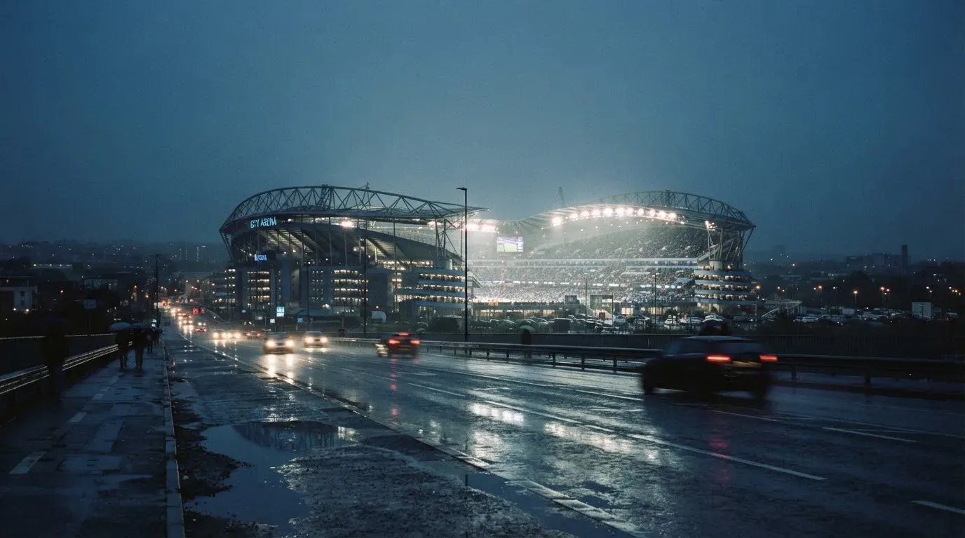 Ein Stadion unter Flutlicht aus großer Entfernung gesehen, davor eine regennasse Straße in der Dämmerung.