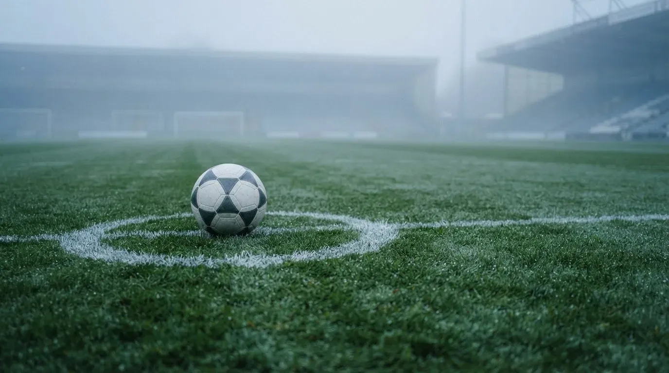 Ein Fußball liegt auf dem Anstoßpunkt eines leeren Rasenplatzes im Morgennebel.