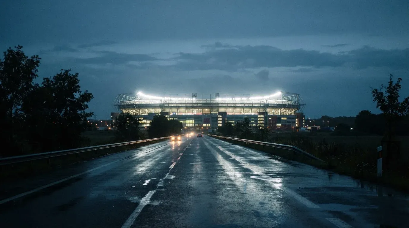 Ein Stadion unter Flutlicht aus großer Entfernung gesehen, davor eine regennasse Straße in der Dämmerung.