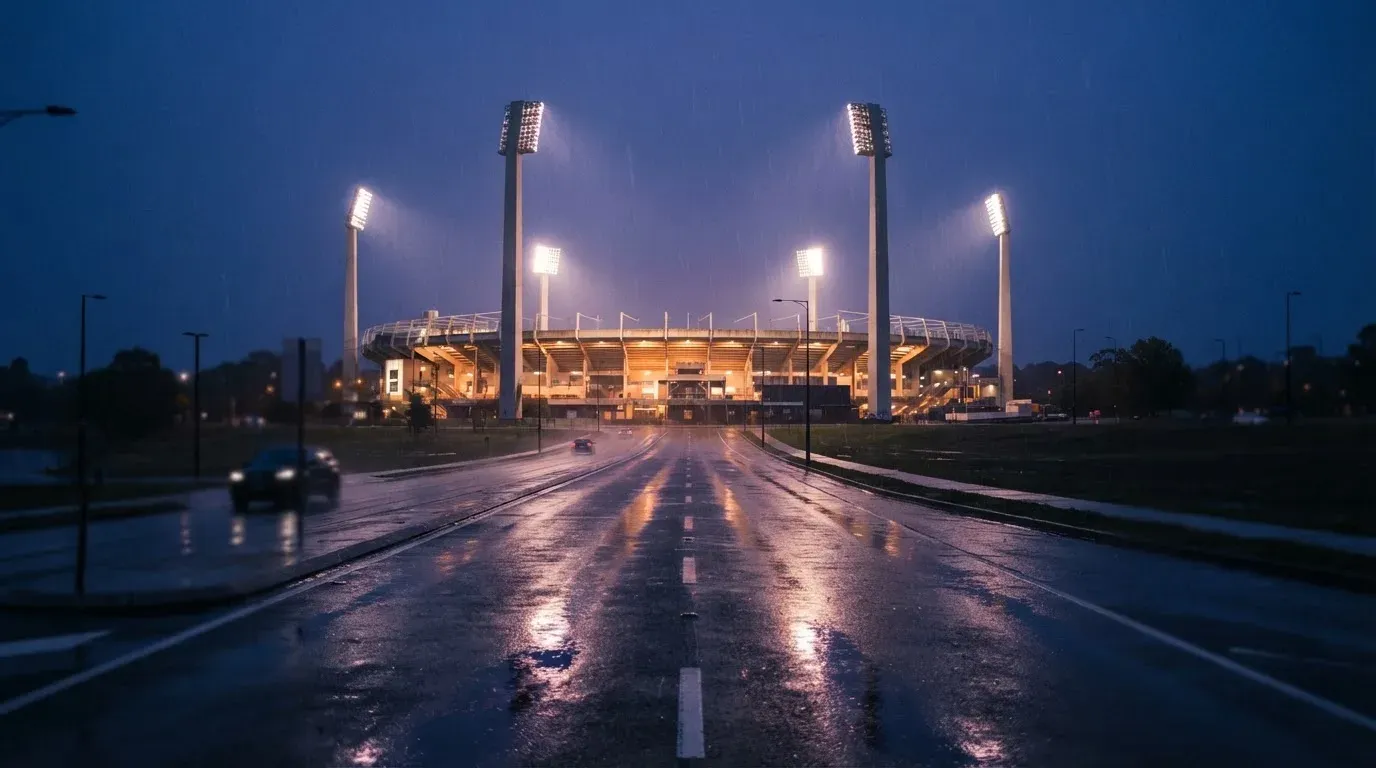 Ein Stadion unter Flutlicht aus großer Entfernung gesehen, davor eine regennasse Straße in der Dämmerung.
