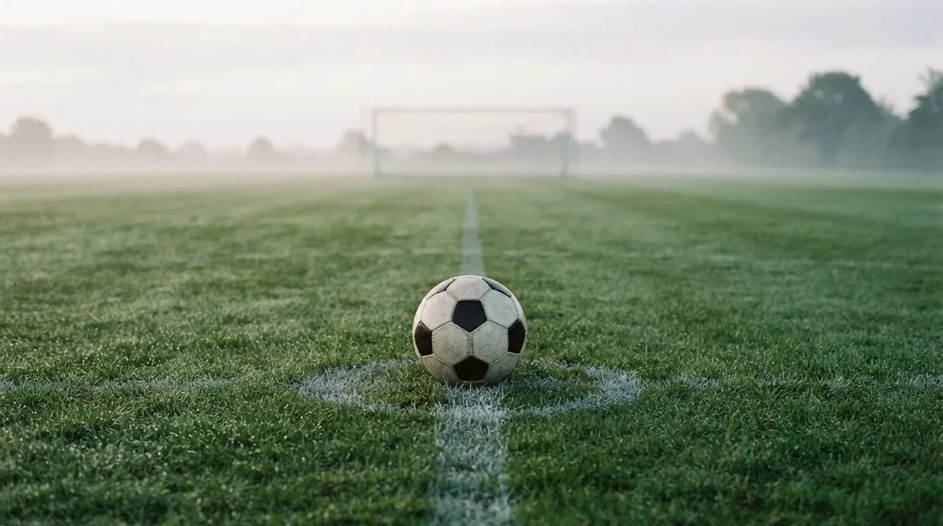 Ein Fußball liegt auf dem Anstoßpunkt eines leeren Rasenplatzes im Morgennebel.