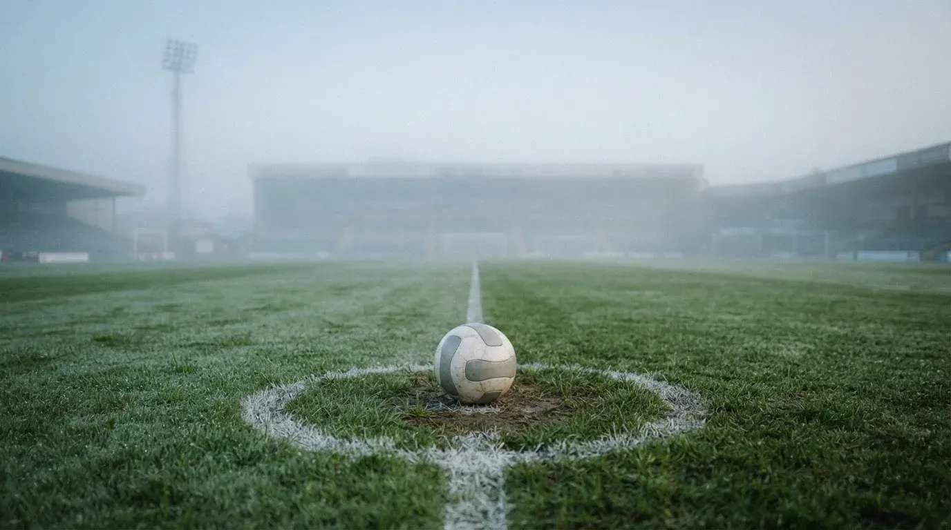 Ein Fußball liegt auf dem Anstoßpunkt eines leeren Rasenplatzes im Morgennebel.