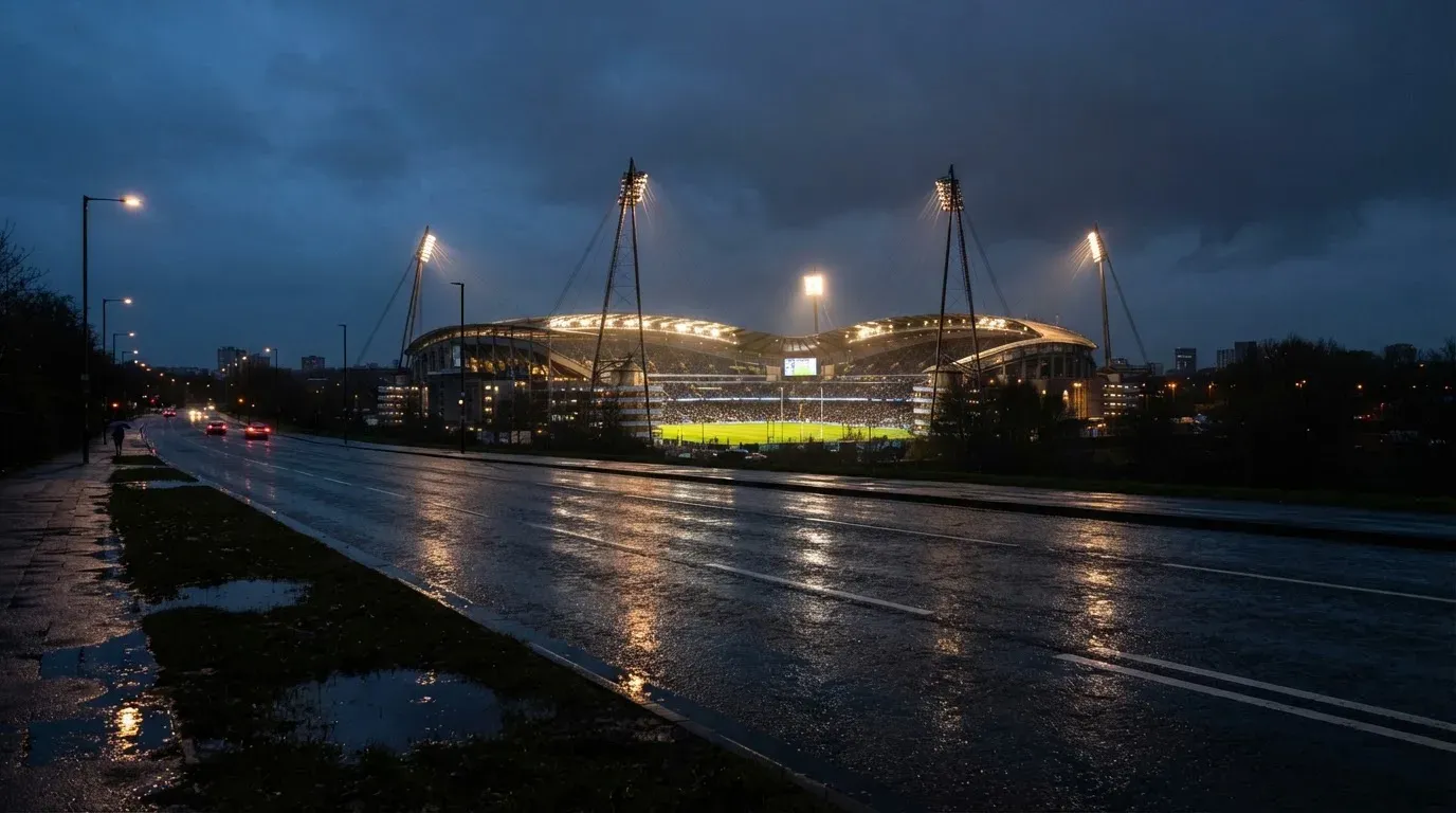 Ein Stadion unter Flutlicht aus großer Entfernung gesehen, davor eine regennasse Straße in der Dämmerung.