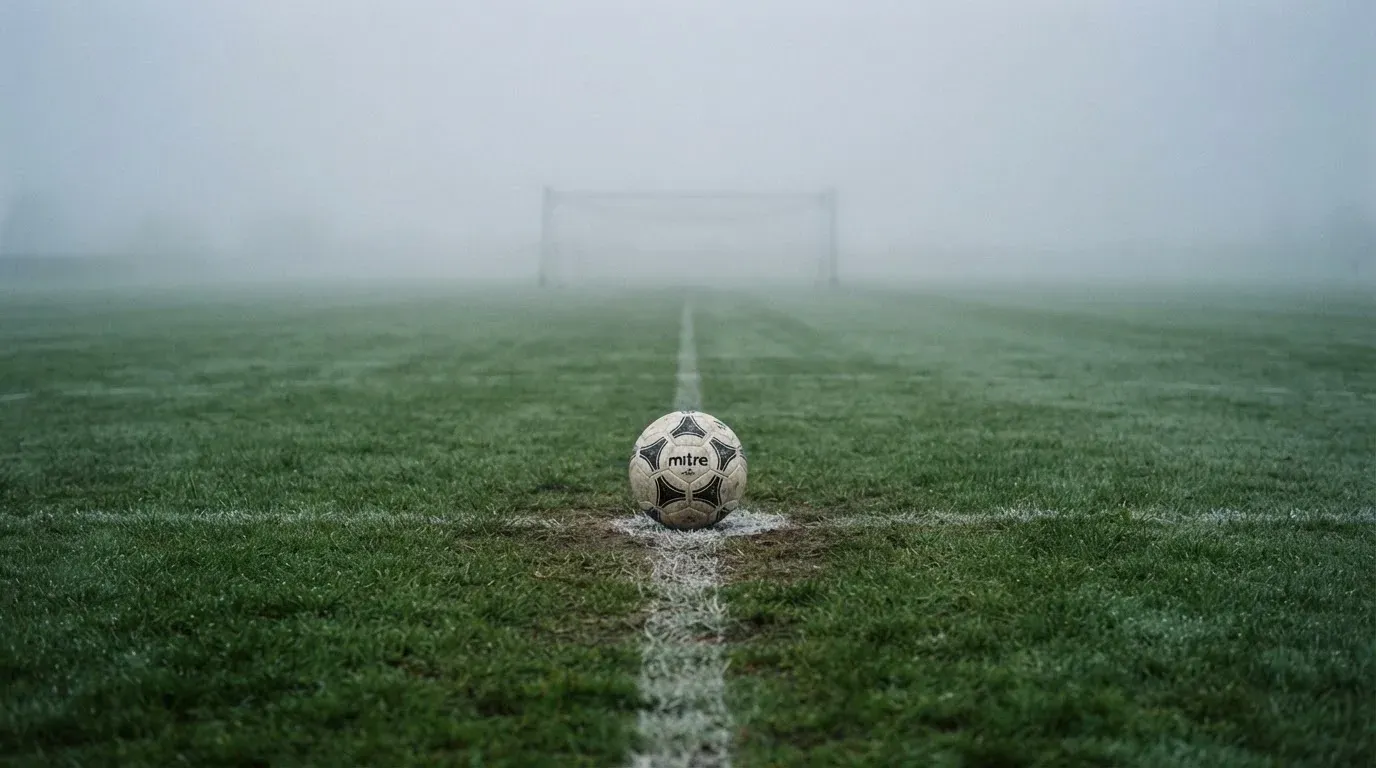 Ein Fußball liegt auf dem Anstoßpunkt eines leeren Rasenplatzes im Morgennebel.