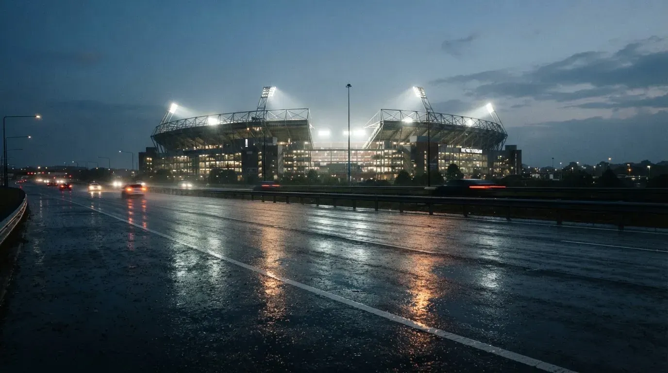 Ein Stadion unter Flutlicht aus großer Entfernung gesehen, davor eine regennasse Straße in der Dämmerung.