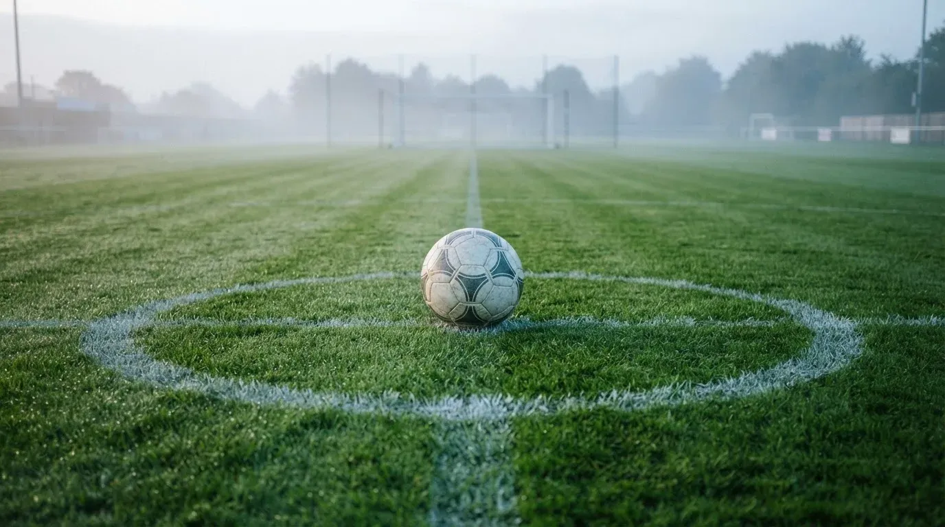 Ein Fußball liegt auf dem Anstoßpunkt eines leeren Rasenplatzes im Morgennebel.
