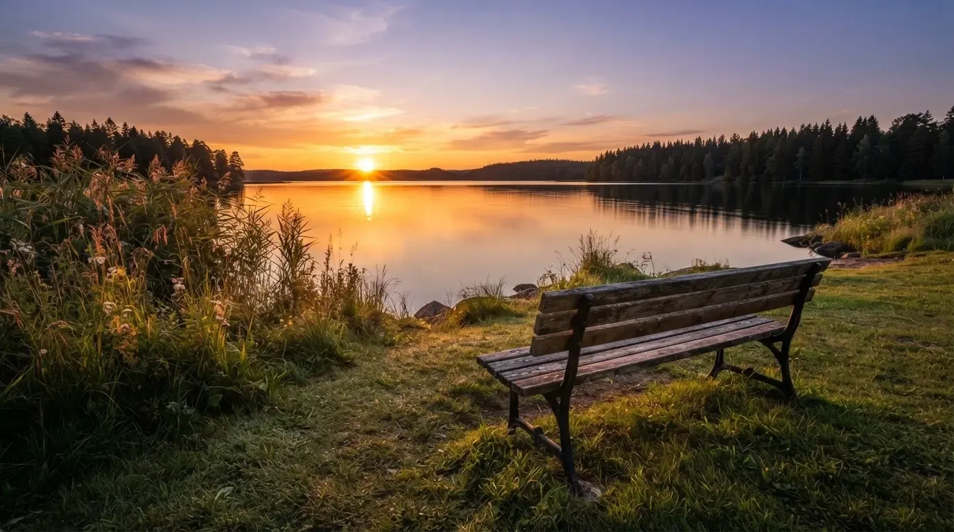 Eine leere Parkbank steht an einem ruhigen See, die Abendsonne spiegelt sich auf dem Wasser.