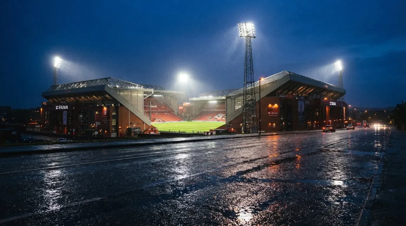 Ein Stadion unter Flutlicht aus großer Entfernung gesehen, davor eine regennasse Straße in der Dämmerung.