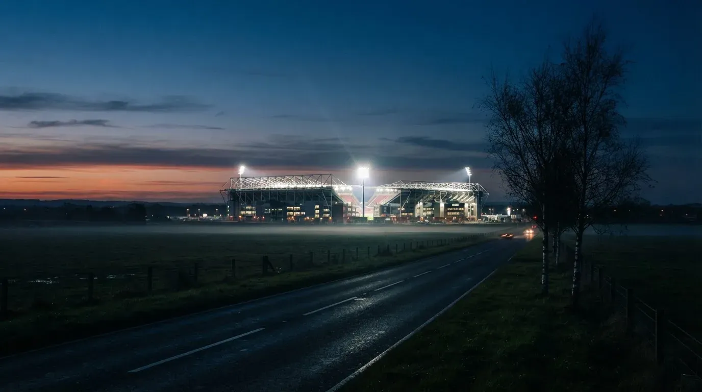 Ein Stadion unter Flutlicht aus großer Entfernung gesehen, davor eine dunkle Landstraße und einzelne Bäume in der Dämmerung.