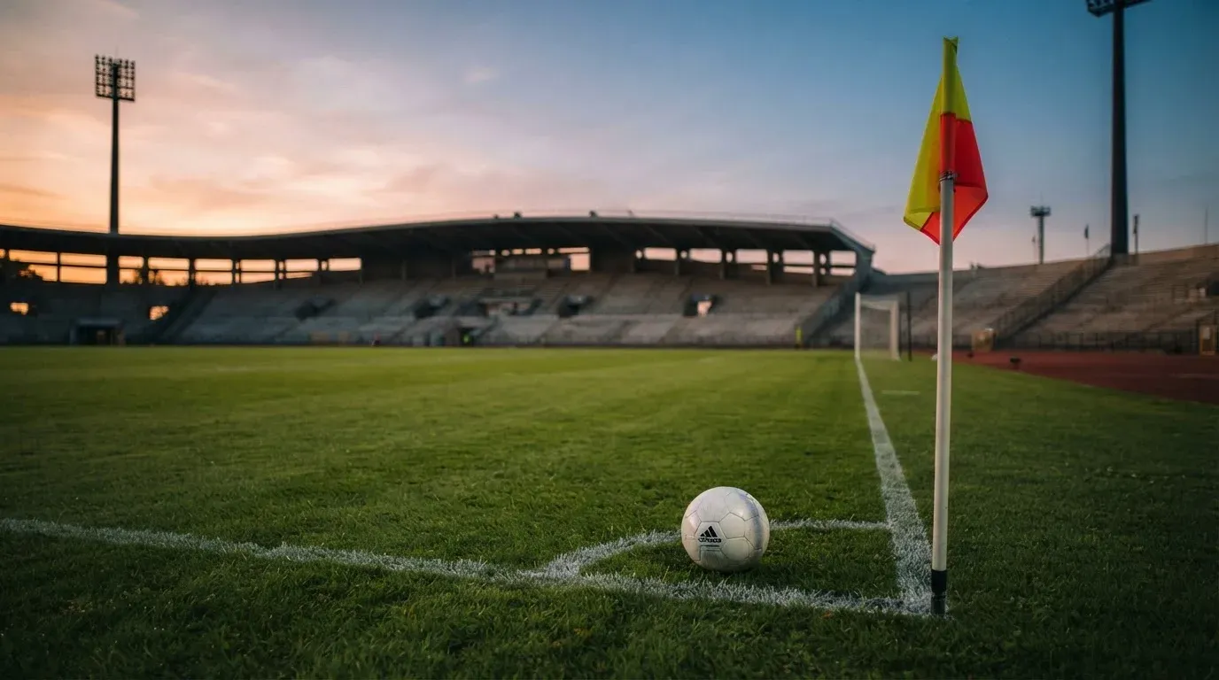 Ein Fußball liegt auf dem Rasen neben der Eckfahne eines leeren Platzes, dahinter eine Tribüne im Abendlicht.