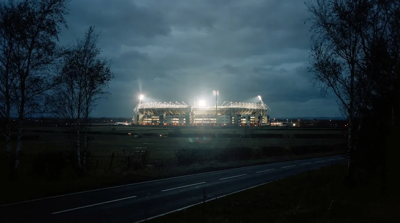Ein Stadion unter Flutlicht aus großer Entfernung gesehen, davor eine dunkle Landstraße und einzelne Bäume.