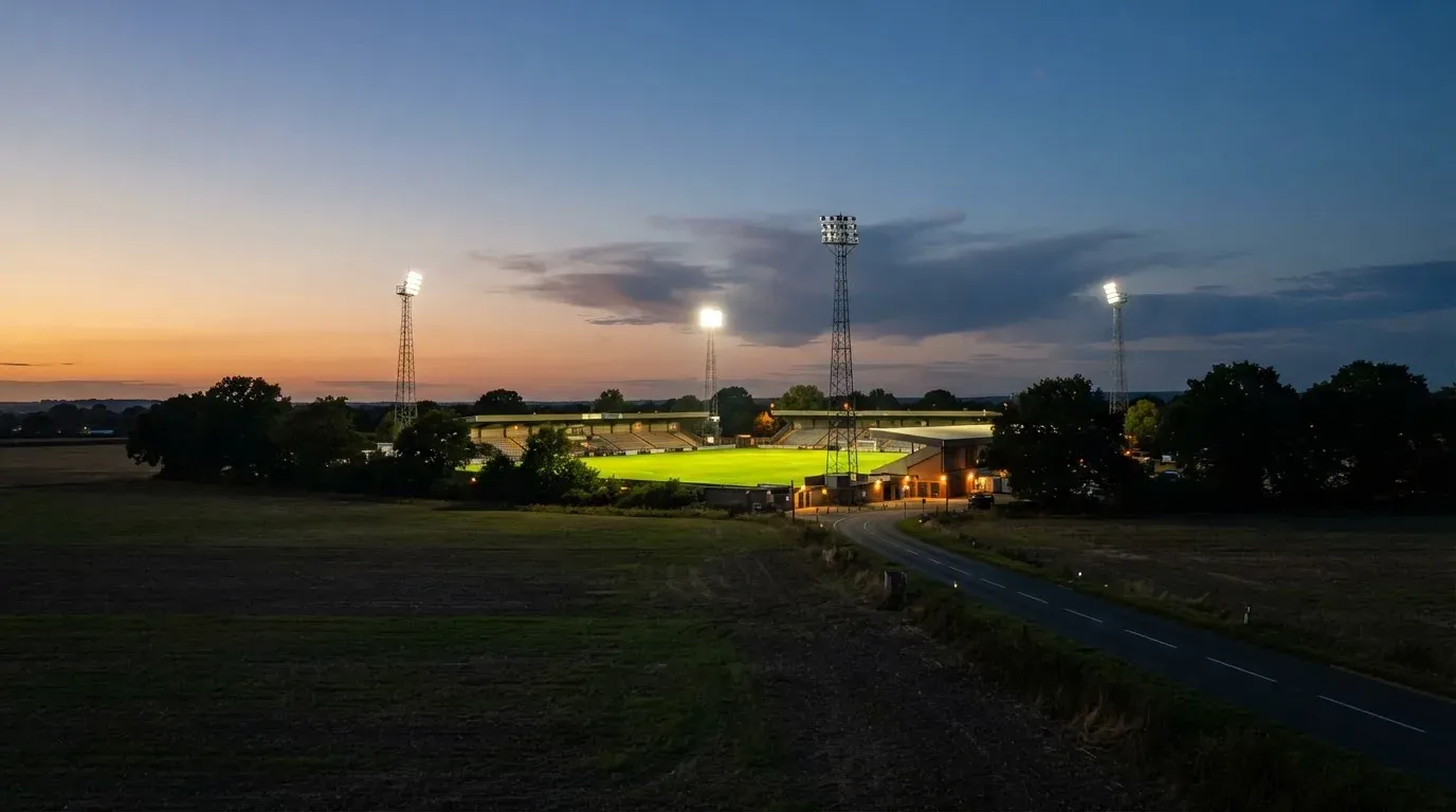 Ein Fußballstadion unter Flutlicht aus großer Entfernung gesehen, davor Felder und eine ruhige Straße in der Dämmerung.