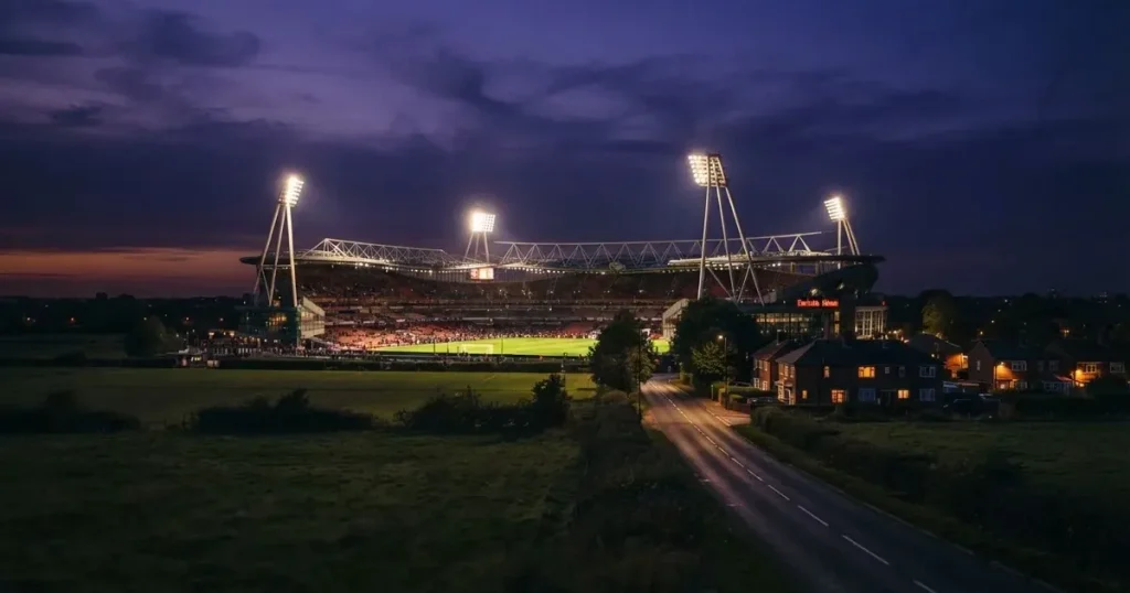 Ein Fußballstadion unter Flutlicht aus großer Entfernung gesehen, davor Felder und eine ruhige Straße in der Dämmerung.