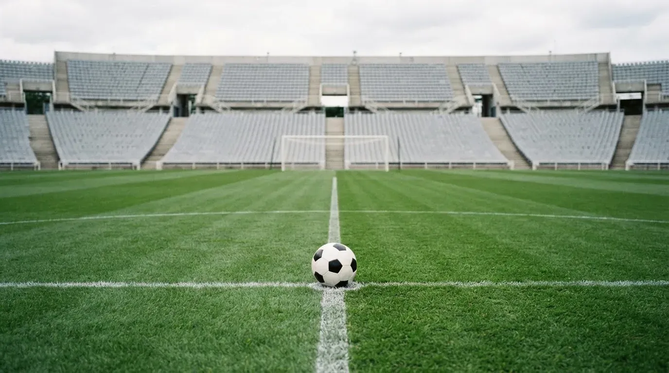 Ein Fußball liegt auf dem Anstoßpunkt eines leeren Rasenplatzes, dahinter eine leere Tribüne in weichem Licht.