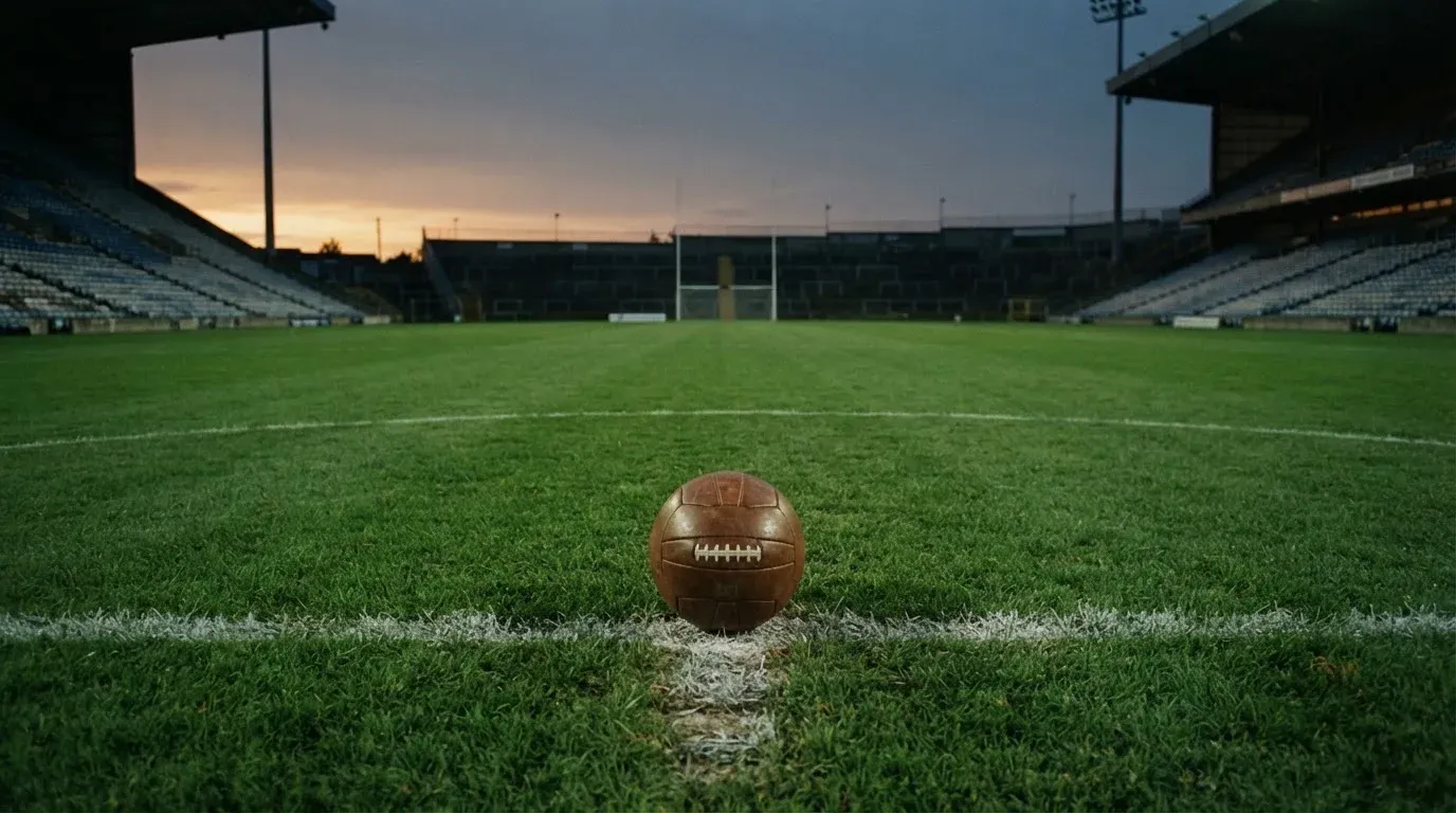 Ein Fußball liegt auf dem Anstoßpunkt eines leeren Rasenplatzes, im Hintergrund eine menschenleere Tribüne im weichen Abendlicht.