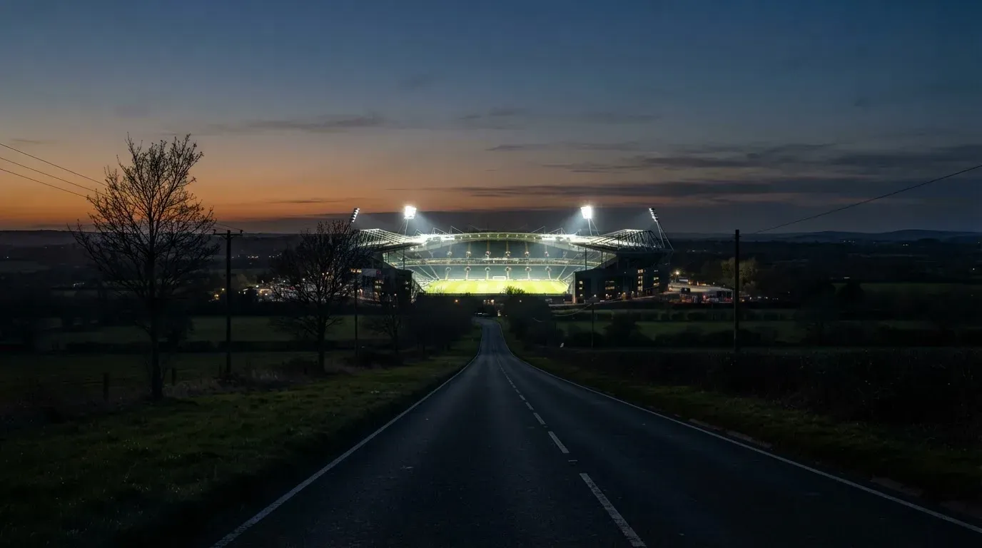 Ein Stadion unter Flutlicht aus großer Entfernung gesehen, davor eine dunkle Landstraße und einzelne Bäume in der Dämmerung.