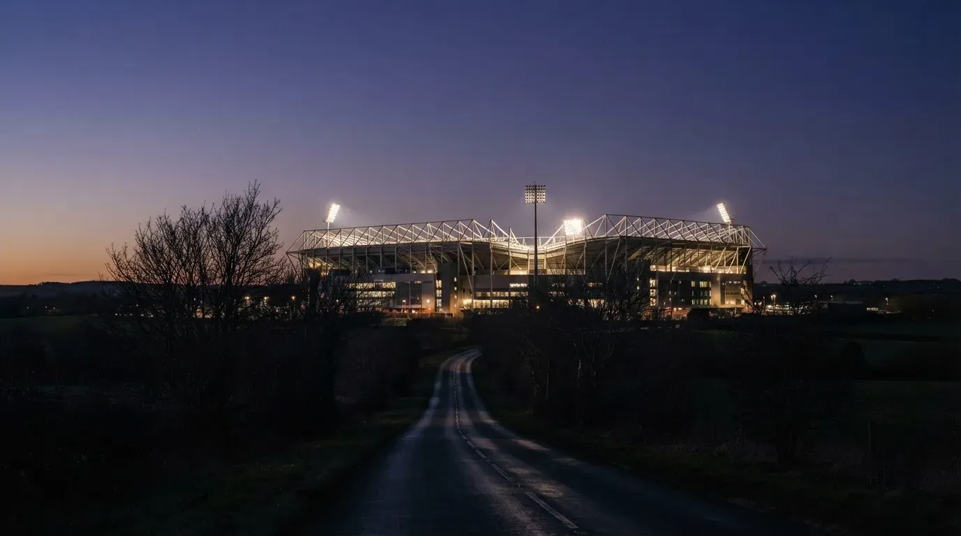 Ein Stadion unter Flutlicht aus großer Entfernung gesehen, davor eine dunkle Landstraße und einzelne Bäume in der Dämmerung.