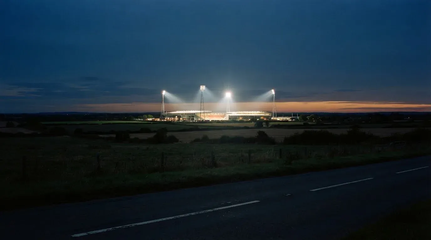 Ein Fußballstadion unter Flutlicht aus großer Entfernung gesehen, davor Felder und eine ruhige Straße in der Dämmerung.