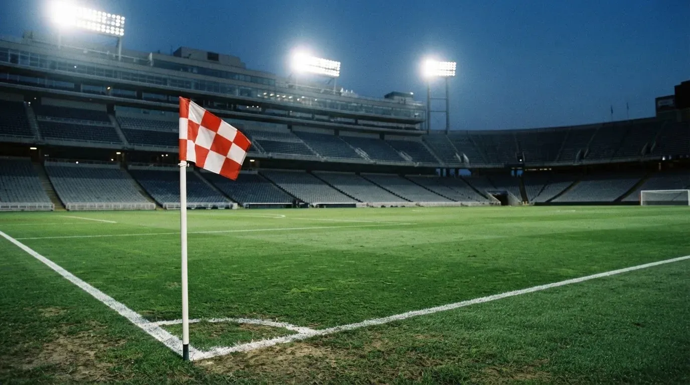 Eine Eckfahne am Rand eines Rasenplatzes, dahinter ein leeres Stadion unter Flutlicht aus der Ferne gesehen.