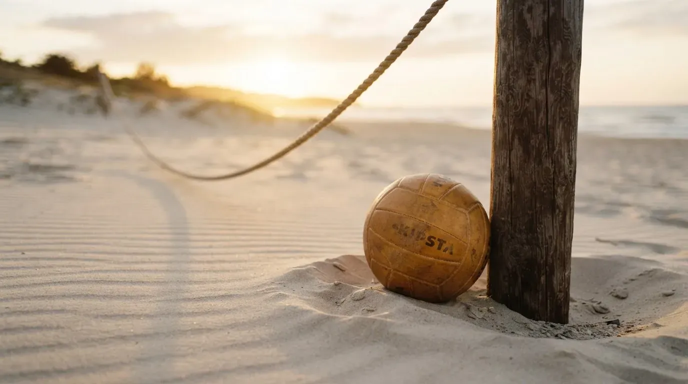 Ein Volleyball liegt auf Sand neben einem Netzpfosten, Strandlicht am späten Nachmittag, im Hintergrund unscharfe Dünen.
