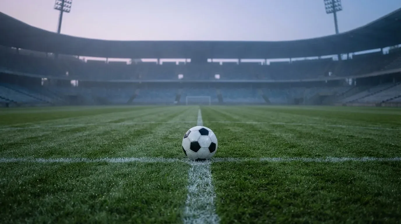 Ein Fußball auf dem Mittelpunkt eines Rasenplatzes, früher Morgen, Tau auf dem Gras, im Hintergrund leere Tribünen.