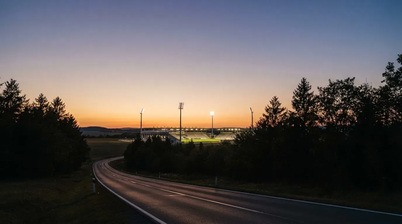 Ein Flutlichtstadion in der Dämmerung aus weiter Ferne, davor ein Wald und eine leere Straße.