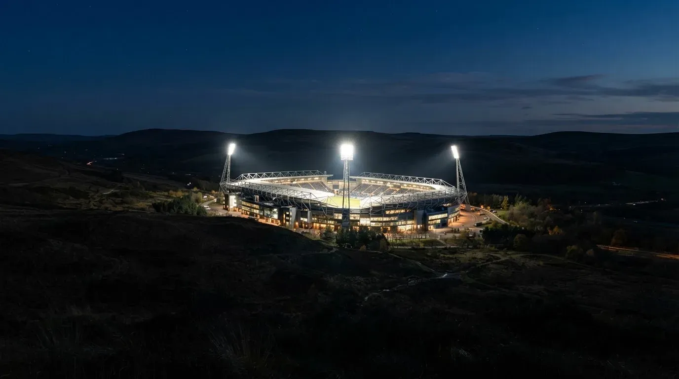 Ein Stadion unter Flutlicht aus großer Entfernung gesehen, darüber ein klarer Nachthimmel und davor dunkle Hügel.