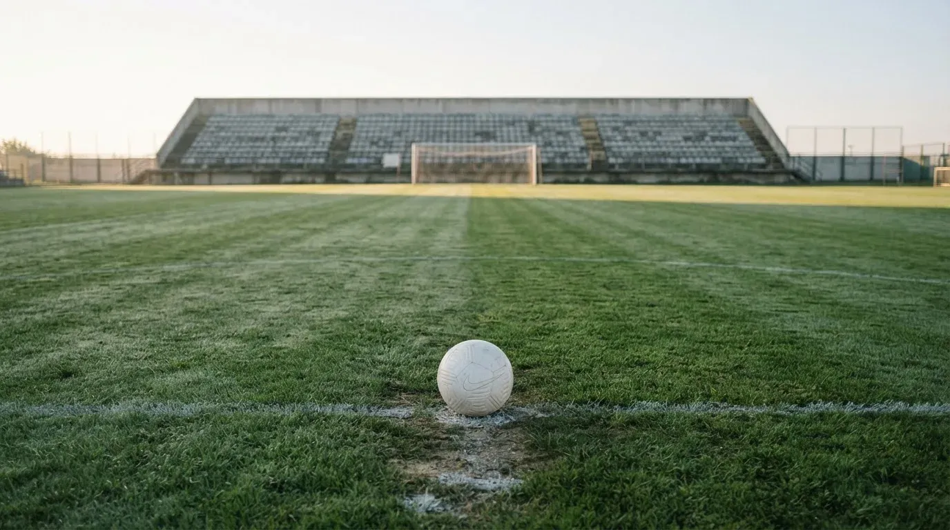 Ein Fußball liegt auf dem Elfmeterpunkt eines leeren Rasenplatzes im Morgenlicht, dahinter eine ruhige Tribüne ohne Zuschauer.