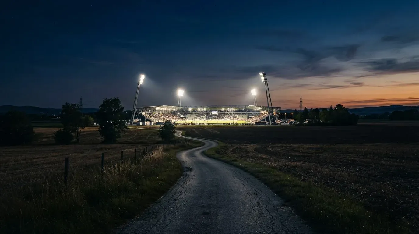 Ein Stadion unter Flutlicht aus der Ferne gesehen, davor eine dunkle Landschaft mit Feldern und einer Landstraße.