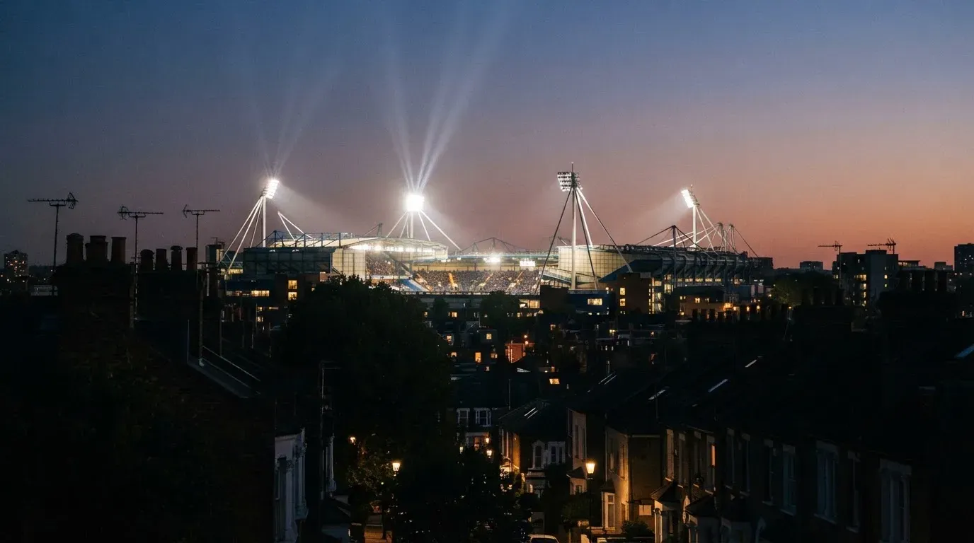 Ein Flutlichtstadion bei Dämmerung aus weiter Ferne gesehen, die Lichtmasten leuchten über Dächern und Bäumen.