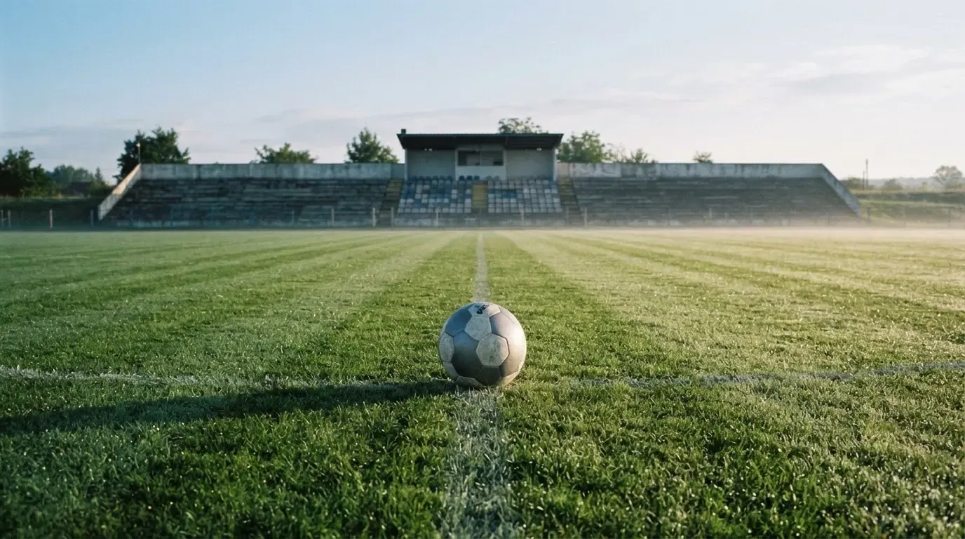 Ein Fußball liegt auf einem Rasenplatz im Morgenlicht, dahinter eine ruhige Tribüne ohne Zuschauer.