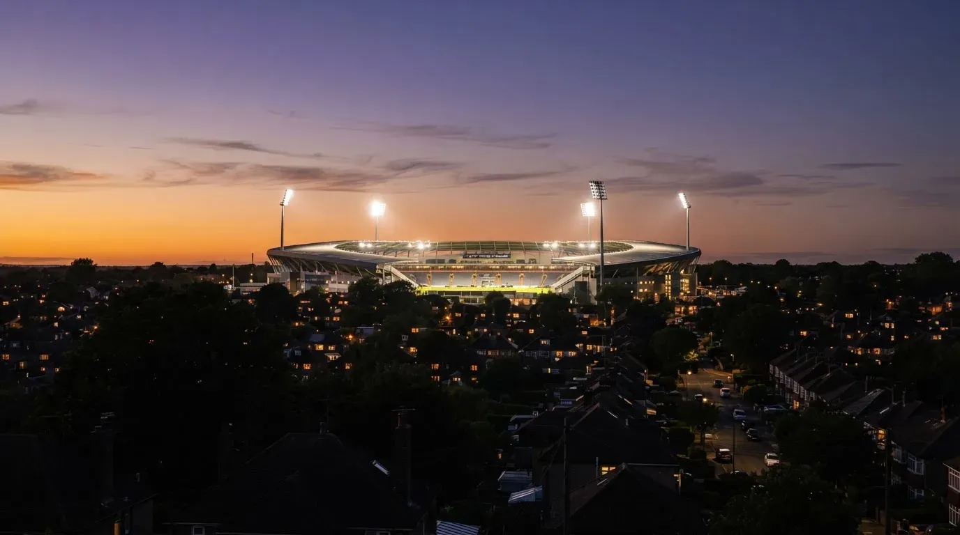 Ein Flutlichtstadion bei Dämmerung aus weiter Ferne gesehen, die Lichtmasten leuchten über Dächern und Bäumen.